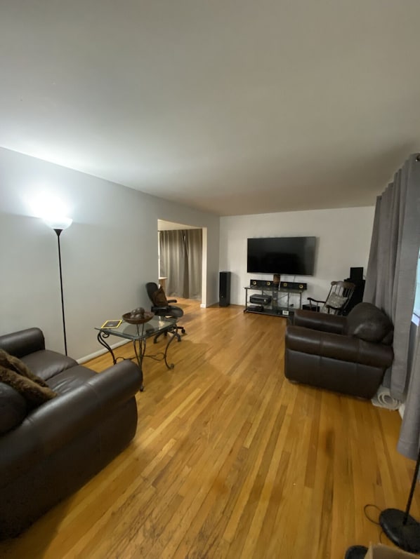 Uma sala de estar espaçosa com piso de madeira, dois sofás de couro, uma mesa de centro de vidro, uma televisão e um sistema de som.