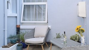 A small outdoor seating area with a wicker chair and table, surrounded by potted plants and a window with blinds. - 2 bedroom accommodation in Sandgate (Sandgate)
