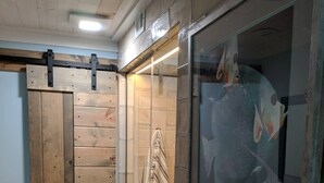 Une salle de bain avec une porte en bois, une douche avec une porte en verre, un lavabo avec un miroir et un tapis à motifs sur le sol.