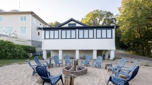 Un espace de détente aménagé sur la plage, avec un foyer, entouré de chaises bleues, devant un bâtiment à deux étages doté d’un toit à pignon.