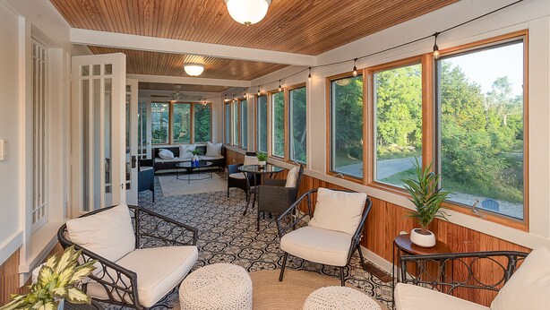 Une véranda avec un plafond en bois, de grandes fenêtres et un coin salon aménagé avec des meubles en osier.