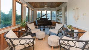 Une pièce avec un grand piano, des fauteuils en osier, des poufs et une vue sur la verdure grâce à de grandes fenêtres.