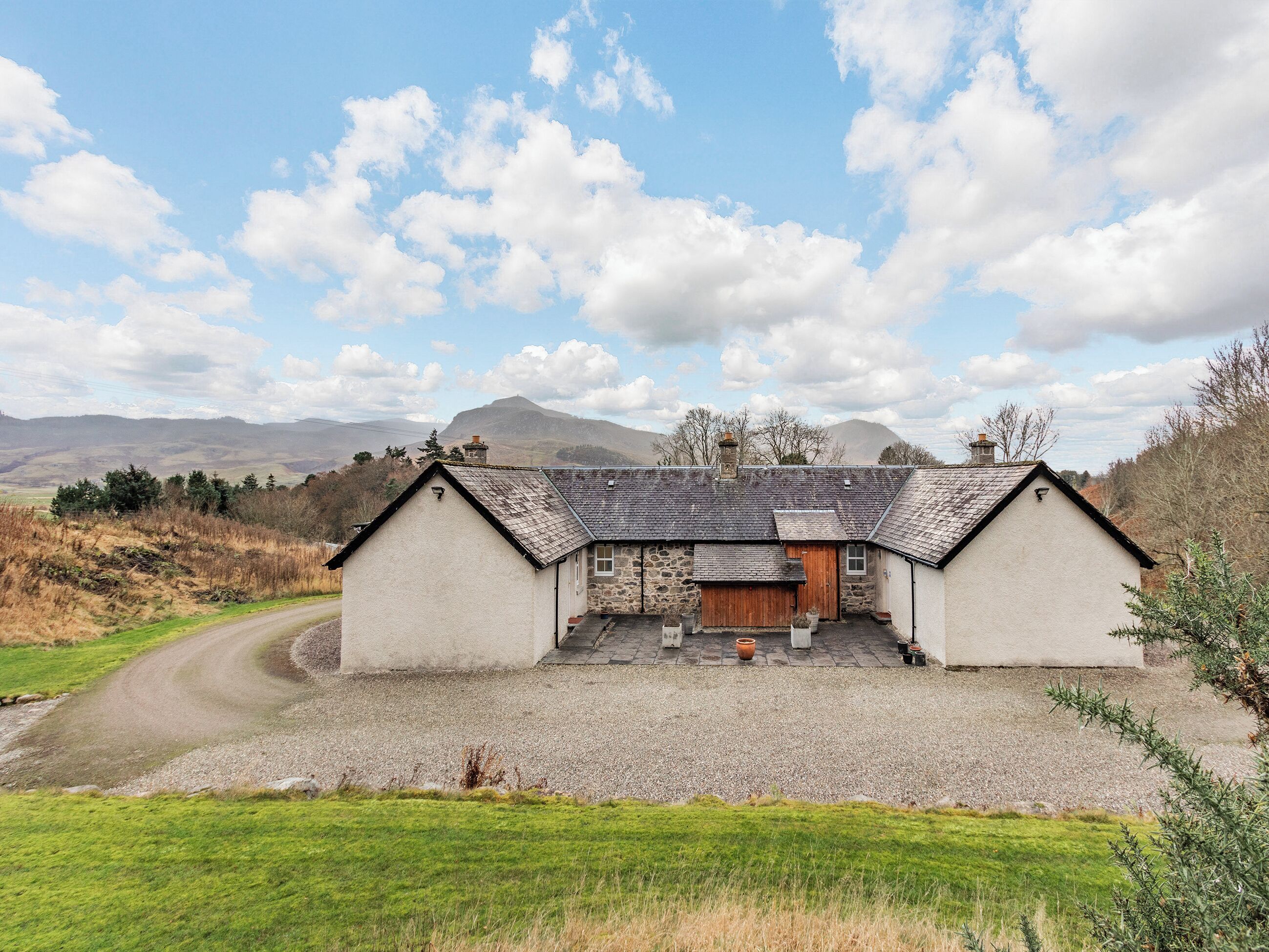 A traditional stone house with a gravel driveway, surrounded by a rural landscape and mountains in the background.