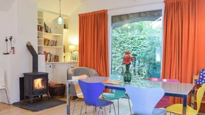 A cozy living room with a wood-burning stove, a dining table with colorful chairs, a bookshelf, and a large window with curtains. - You’ll love the bright open space allowing for some real family fun. (Lothianburn, near Edinburgh)