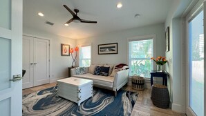 A spacious living room with a ceiling fan, a large window with blinds, a sofa with cushions, a coffee table, and a decorative rug.