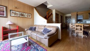 A living room with a brown leather sofa, a glass coffee table, and a red chair. A staircase leads to a kitchen area with wooden cabinets and a dining table. - Monte Solena 45C in Bormio (Bormio)