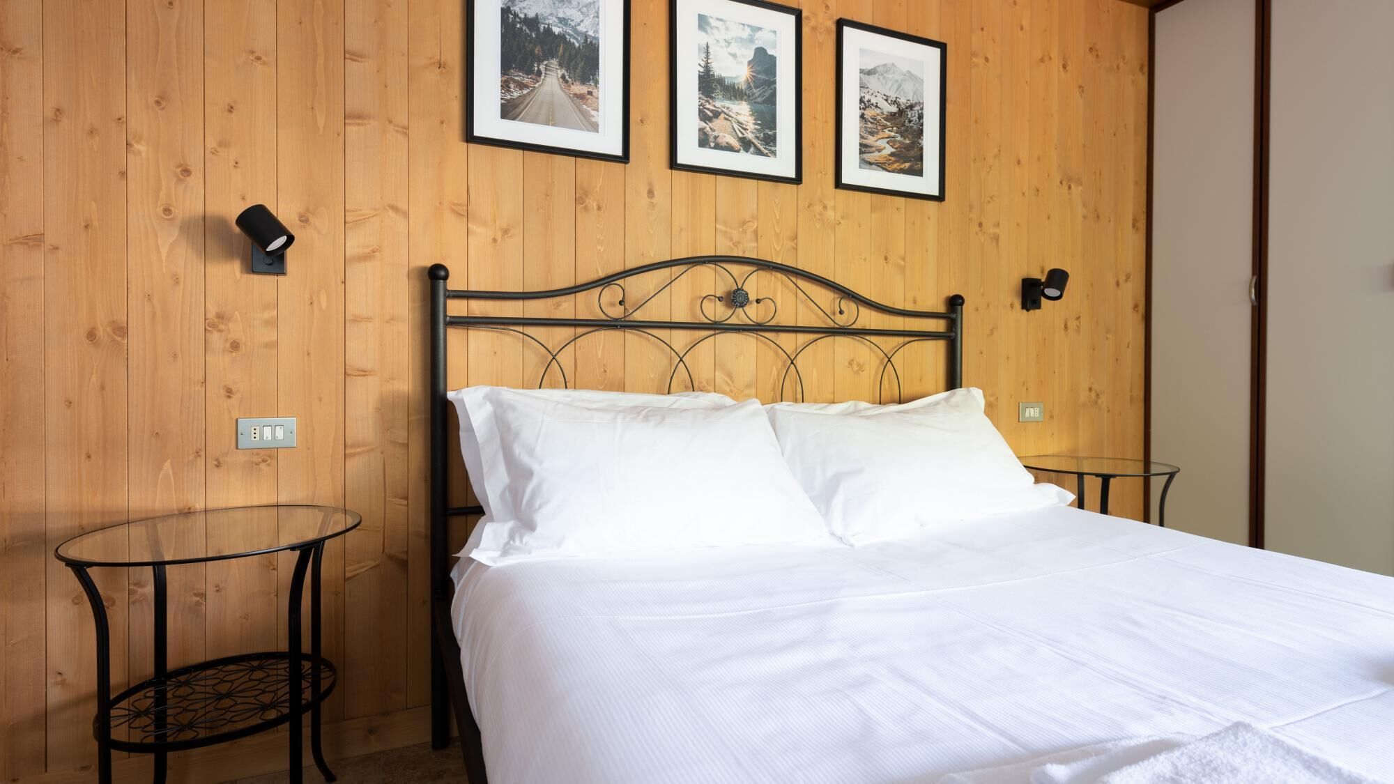 A neatly made bed in a room with wooden panel walls, a small round table, and three framed pictures on the wall.