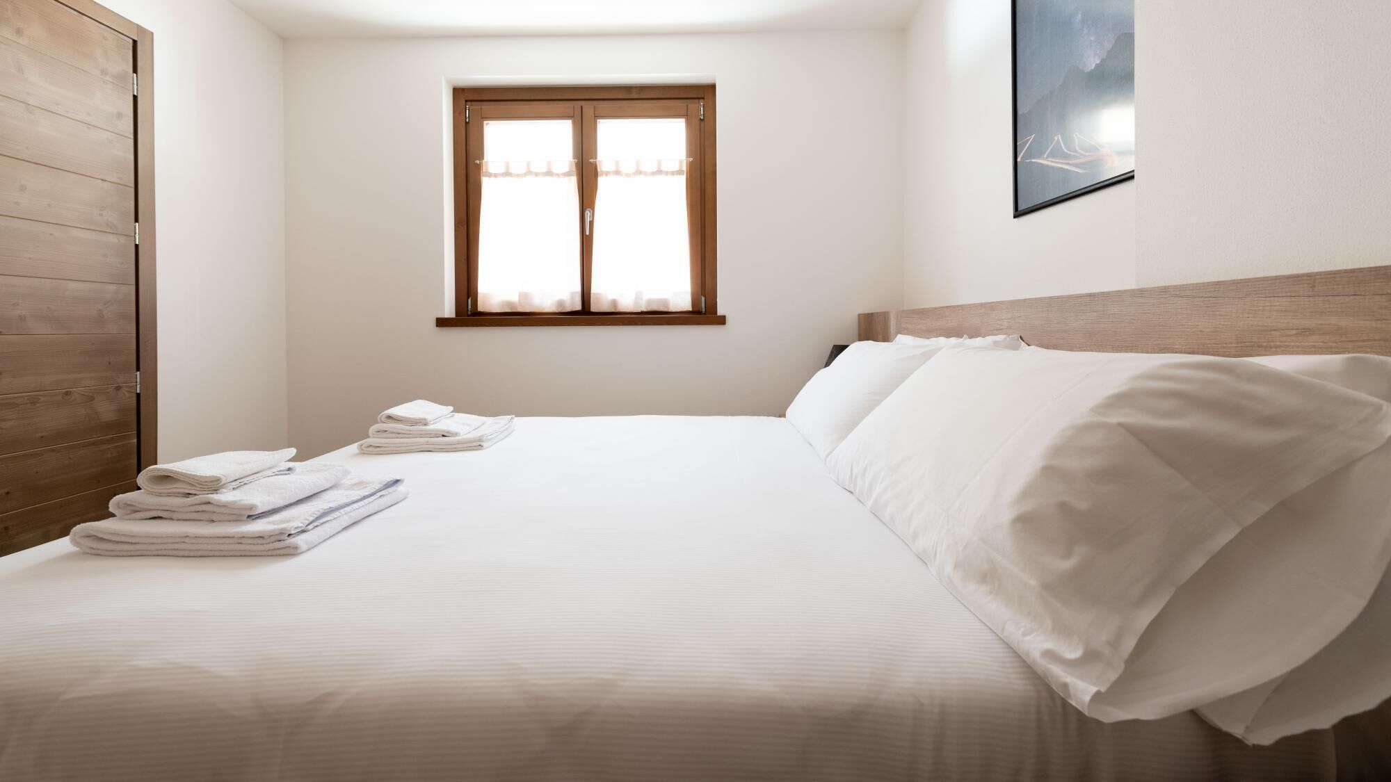 A neatly made bed with white linens and towels, a wooden headboard, a window with sheer curtains, and a framed picture on the wall.