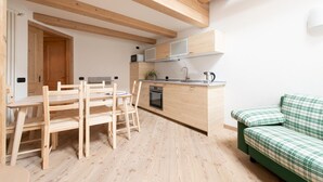 Cocina compacta con armarios de madera, un microondas, un horno y un lavamanos. Hay una zona de comedor con sillas y una mesa de madera. Un sofá verde a cuadros se ve en la esquina.