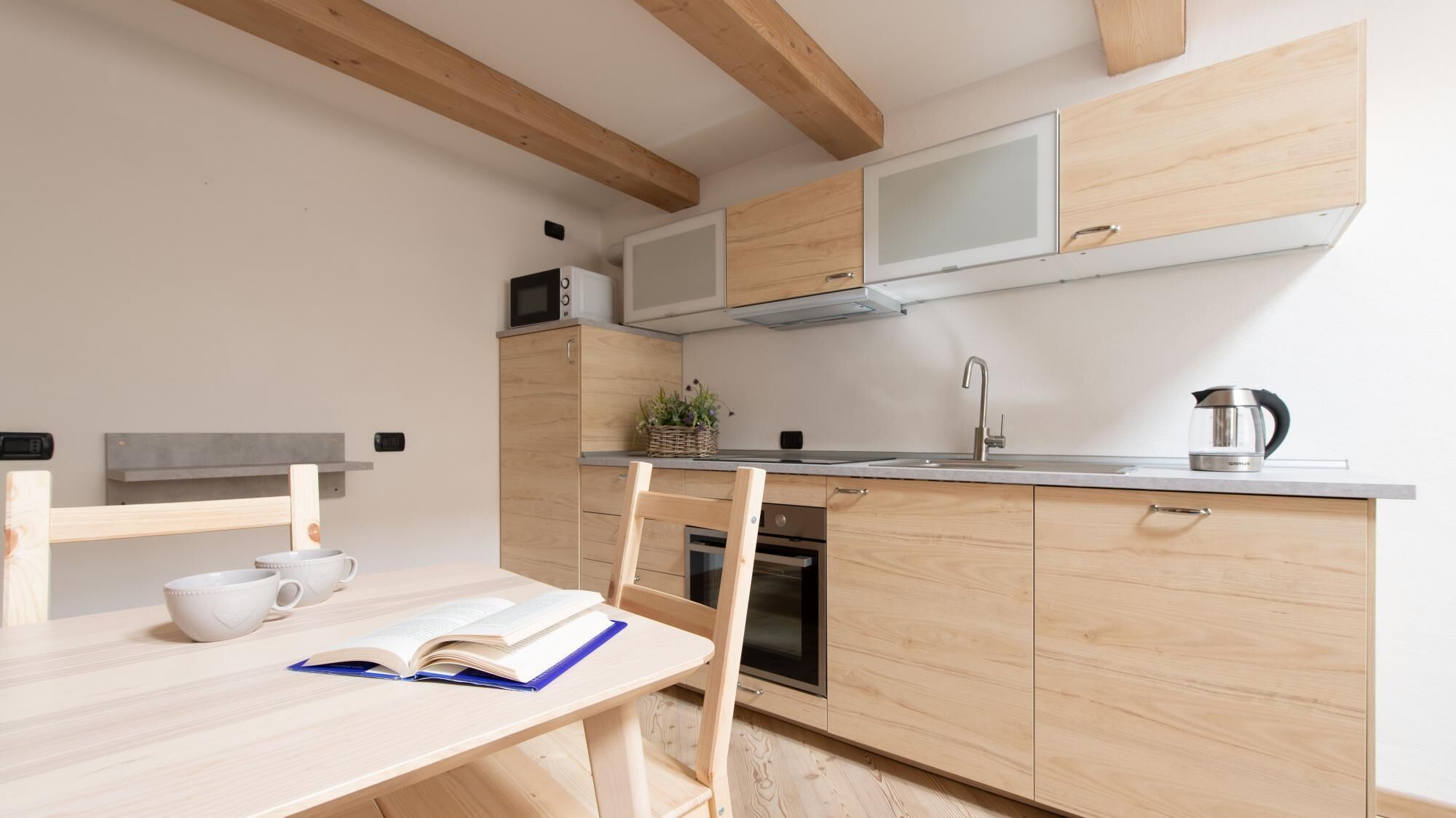 A modern kitchen with wooden cabinets, a microwave, a kettle, and a small dining area with a table and chairs.