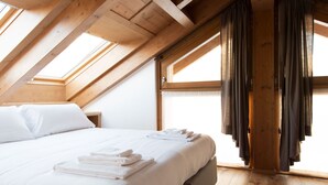 Quarto com teto inclinado, vigas de madeira, cama grande com roupa de cama branca e janela com cortinas.