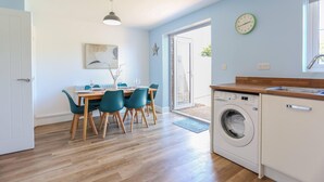 A modern kitchen with a built-in washing machine, a dining area with a table and chairs, and a large window providing natural light. - Elements 3 bed Home in Bracklesham Bay (Chichester)