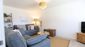 A modern living room with a grey sofa, a round coffee table, a television on a wooden stand, and a hanging light fixture. - Elements 3 bed Home in Bracklesham Bay (Chichester)