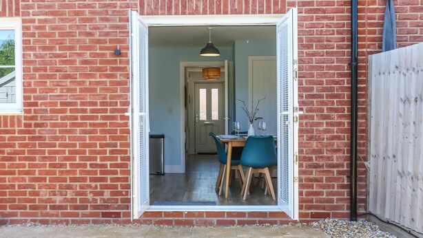 A brick exterior with a white-framed door leading to a dining area with a table, chairs, and a hanging light fixture. - Elements 3 bed Home in Bracklesham Bay (Chichester)