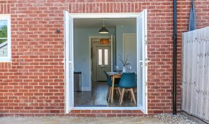 A brick exterior with a white-framed door leading to a dining area with a table, chairs, and a hanging light fixture. - Elements 3 bed Home in Bracklesham Bay (Chichester)