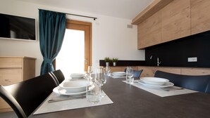 Comedor con una mesa dispuesta para cuatro, incluyendo platos, vasos y cubiertos. Hay sillas alrededor de la mesa, y se ve una cocina al fondo.