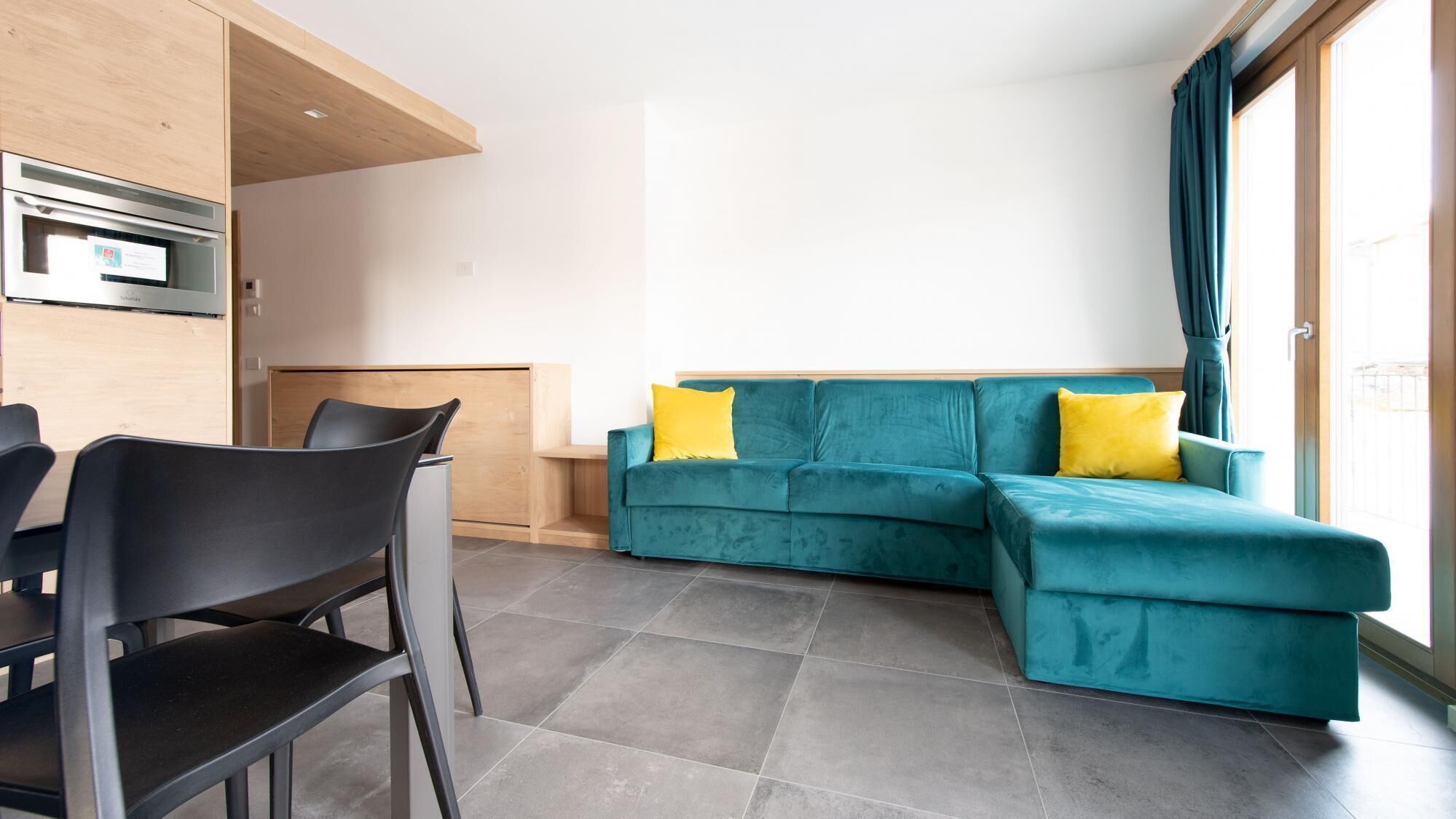 A modern living room with a teal sofa, two yellow cushions, a wooden dining table with chairs, and a built-in oven.