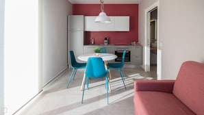 A modern kitchen with white cabinets, a red accent wall, and a dining area with blue chairs and a white table. - Villa Paloma - Levante Bs-e967-prov4at (Marone)