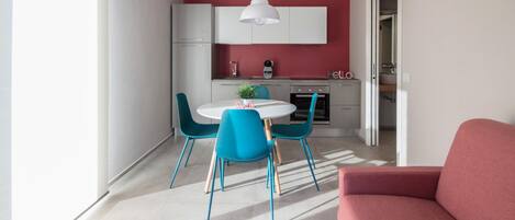 A modern kitchen with white cabinets, a red accent wall, and a dining area with blue chairs and a white table.