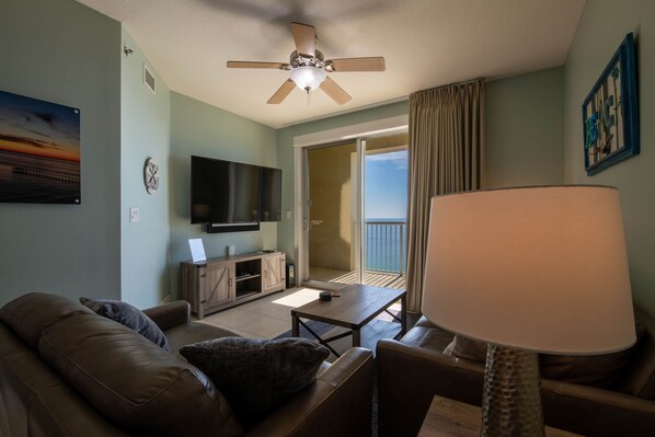 A living room with a brown leather sofa, a wooden coffee table, a TV on the wall, a ceiling fan, and a view of the outdoors through a sliding door.