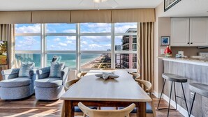 Una sala de estar moderna con un ventanal grande que da a una playa y rascacielos. Hay una mesa de comedor con sillas y una zona de cocina con taburetes de bar.
