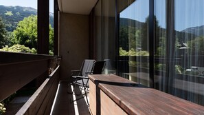 En balkon med et træbord og stole, store vinduer der giver udsigt over et bjergrigt landskab.