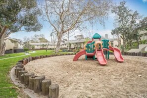 A playground with a slide and climbing structure, surrounded by a sandy area and wooden posts.