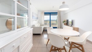 Una sala de estar moderna con un sofá blanco, una mesa de comedor con sillas, un televisor y un ventanal con vistas.