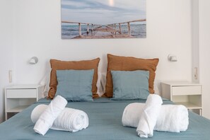 Een net opgemaakt bed met blauwe en bruine kussens, witte nachtkastjes en een schilderij van een pier aan de muur.