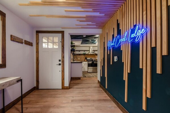 A hallway with a wooden floor, a neon sign reading 'Mud Creek Lodge,' and a door leading to a kitchen area.