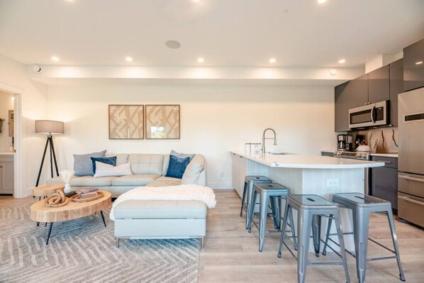 A modern living room with a sofa, a coffee table, and a kitchen area with a sink and bar stools. - FLX Solar Powered Village/Tunnel to Seneca Lake! (Geneva)