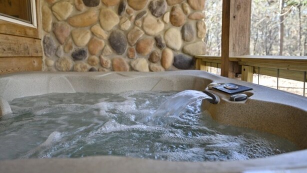 A hot tub with a stone wall backdrop and wooden deck railing. -  Secluded Hochatown Cabin w/ Hot Tub, Fire Pit - Sleeps 6 🌲 (broken bow)