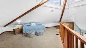 Ein Schlafzimmer mit schräg angesetzter Decke, einem Bett mit gemusterten Kissen, einem kleinen Tisch mit Lampe und einer Holzhandläufe.
