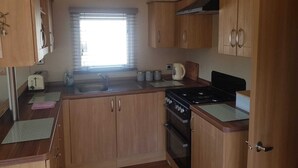 A compact kitchen with wooden cabinets, a gas stove, a sink, and a window with blinds. - Modern 6 berth caravan close to fishing lakes and beach (Burnham-on-Sea)