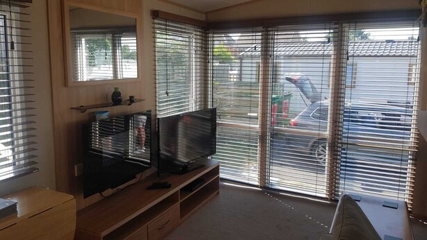 A living room with a TV, wooden furniture, and large windows with blinds. - Modern 6 berth caravan close to fishing lakes and beach (Burnham-on-Sea)