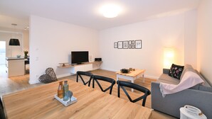 Ein modernes Wohnzimmer mit einem Holzesstisch, schwarzen Stühlen, einem grauen Sofa, einem Flachbildfernseher und einem Wandregal mit gerahmten Bildern.
