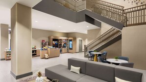 Un hall d’entrée spacieux avec un escalier moderne, un coin salon avec un canapé et des fauteuils, et un stand de vente de nourriture en arrière-plan.