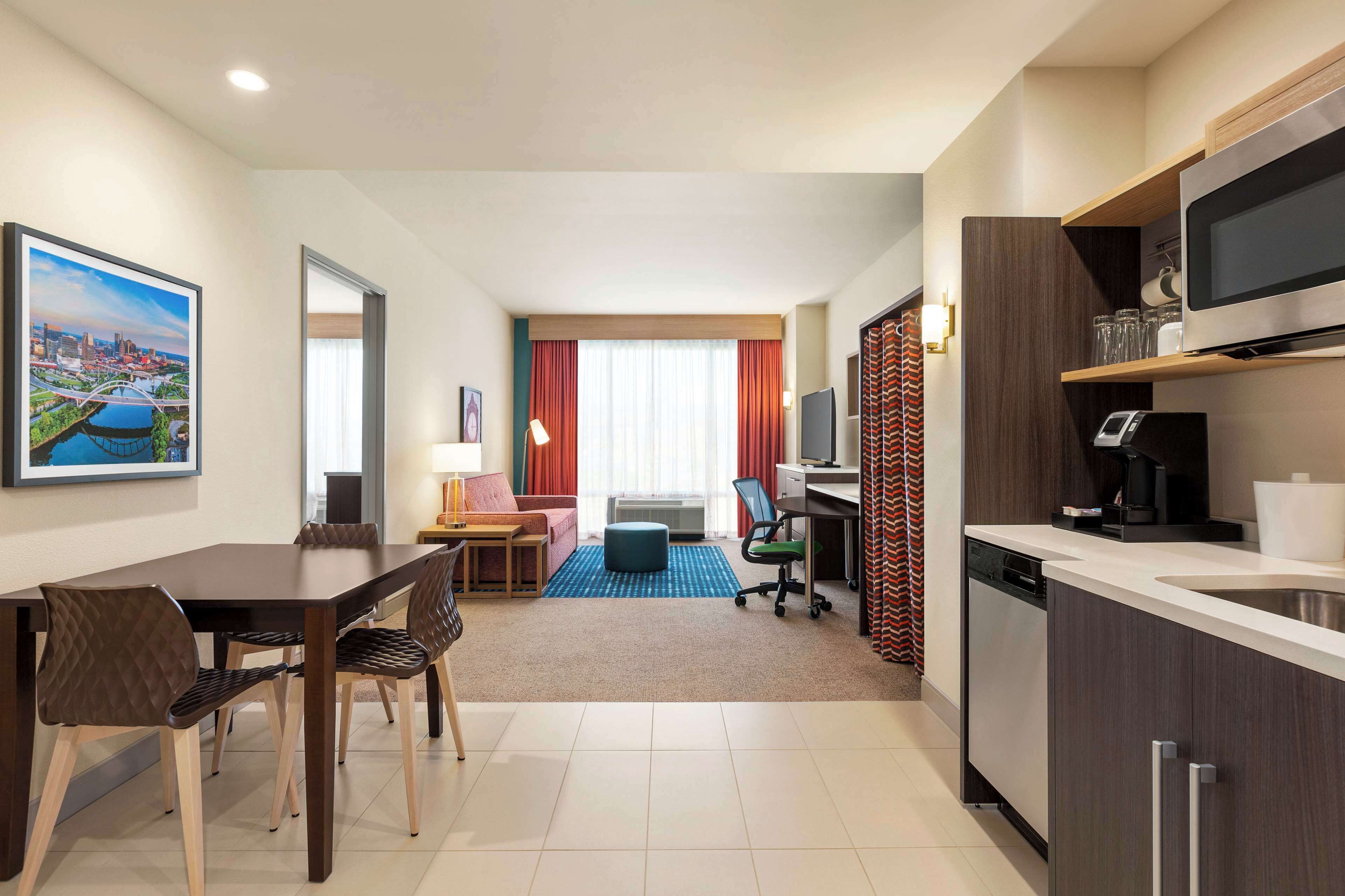 A modern hotel room with a dining area, kitchenette, and a large window with curtains.