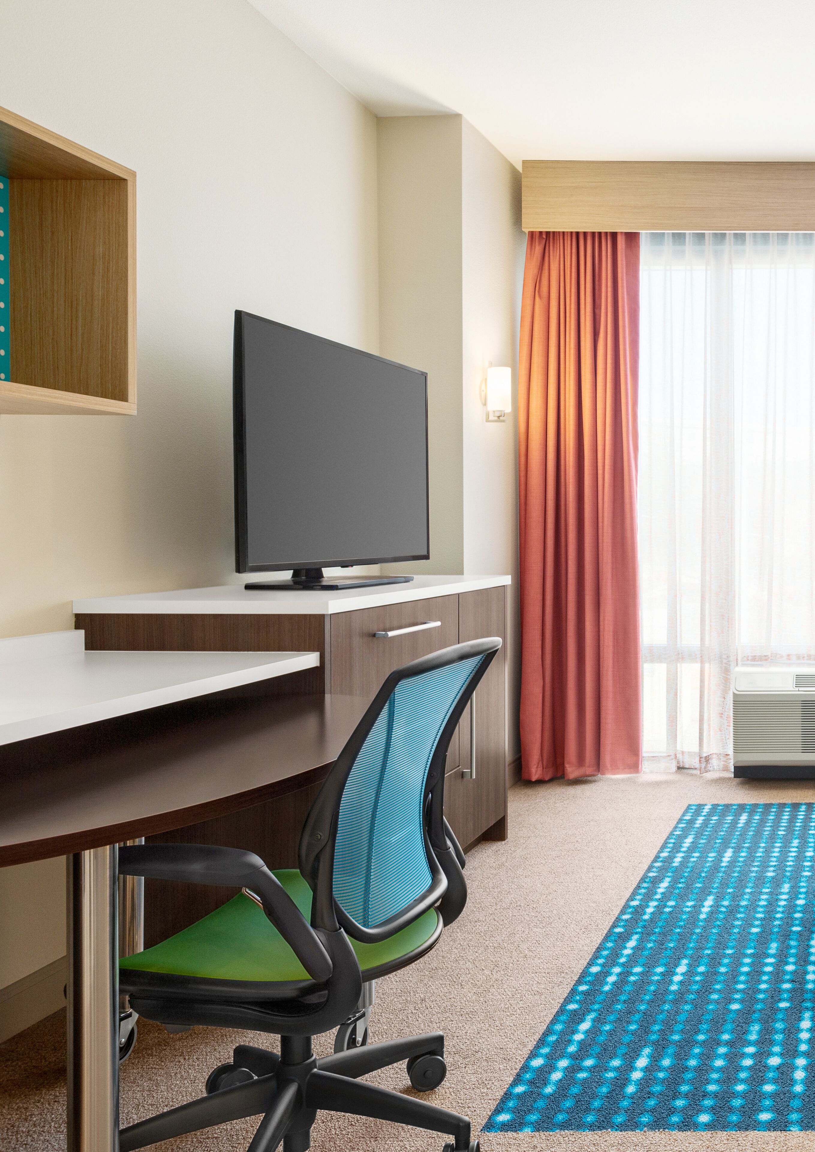 A hotel room with a desk, chair, television, window with curtains, and a carpet.