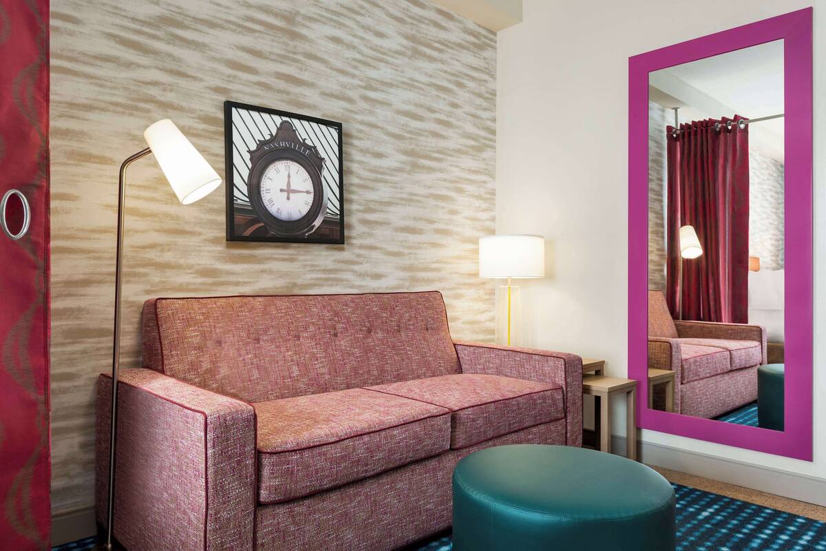 A modern hotel room with a pink sofa, a round teal ottoman, a wall clock, a mirror, and a patterned wall.