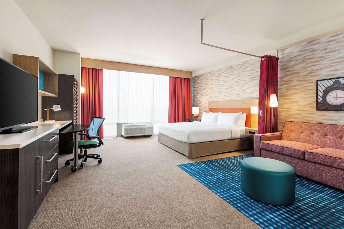 A hotel room with a bed, a desk with a computer, a TV, a sofa, and a large window with red curtains.