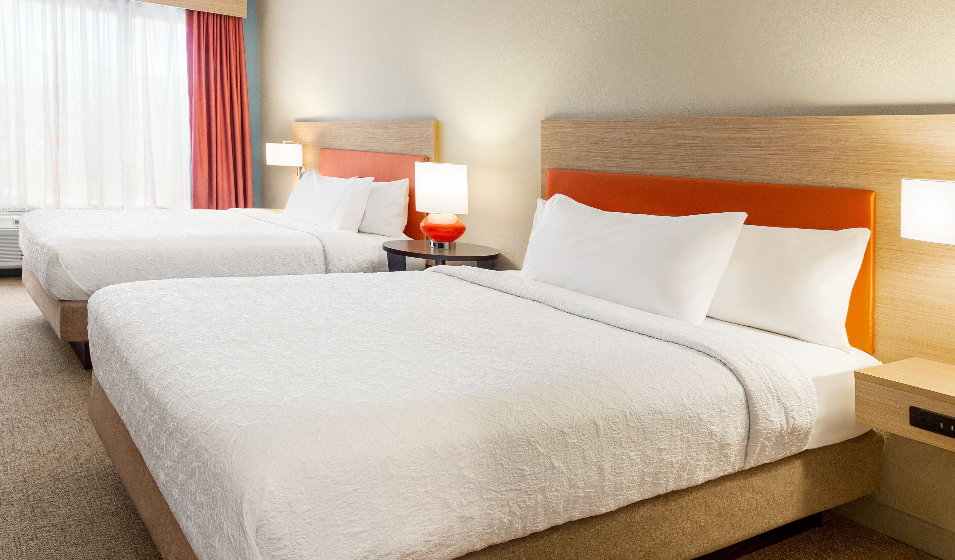 Una habitación de hotel con dos camas, cada una con ropa de cama blanca y un cabecero naranja. Entre las camas hay una mesita de noche con una lámpara.