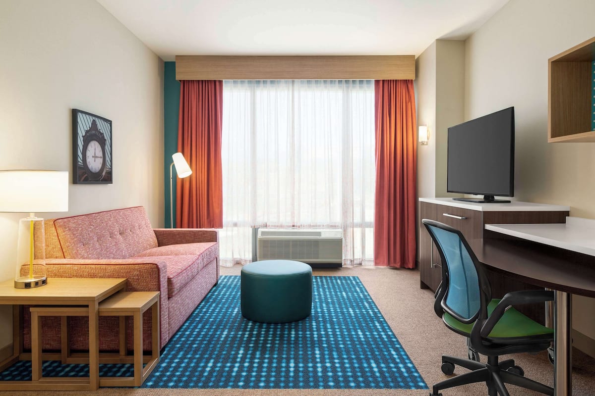 A hotel room with a sofa, ottoman, desk, chair, TV, and air conditioning unit.