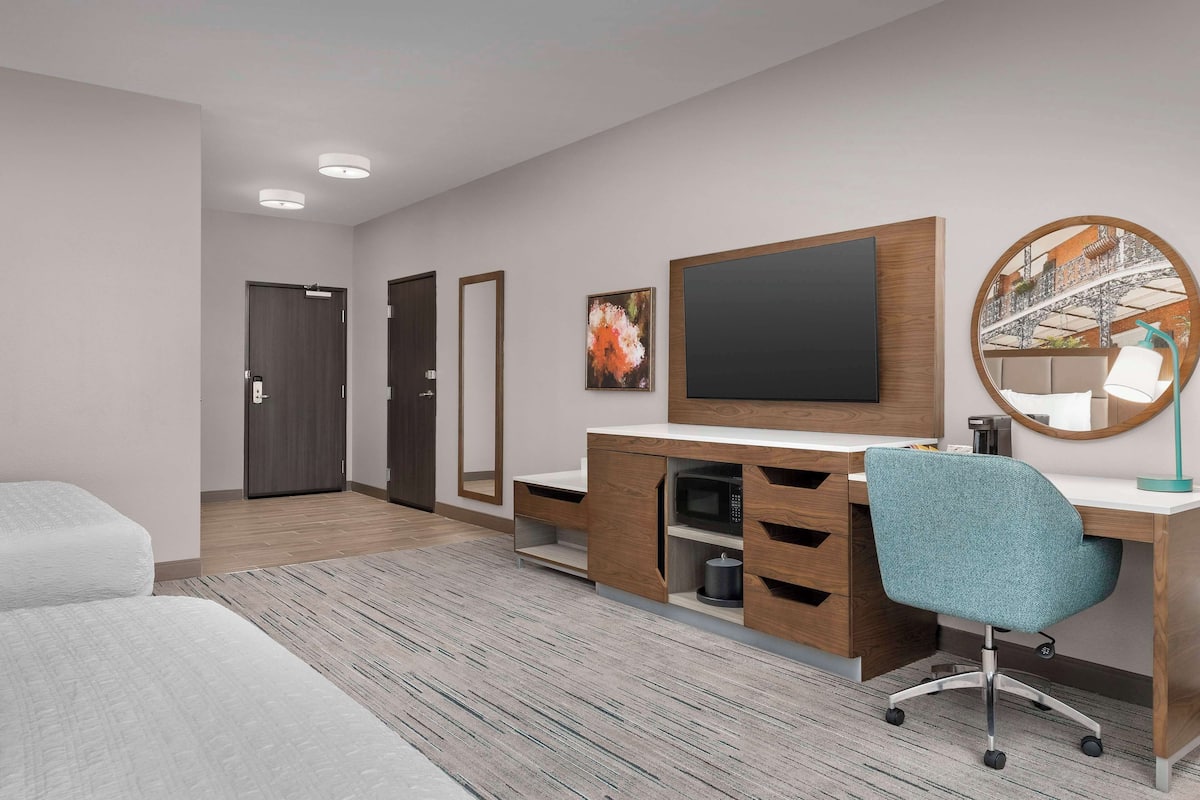 A modern hotel room with a wooden desk, a teal office chair, a flat-screen TV, a round mirror, and a bed.