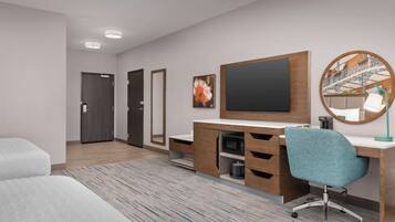 A modern hotel room with a wooden desk, a teal office chair, a flat-screen TV, a round mirror, and a bed.