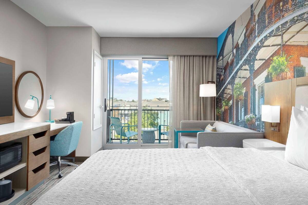 A hotel room with a large bed, a desk with a chair, a small table, a sofa, and a balcony with a view of the city.