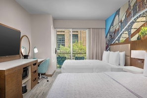 Habitación de hotel con dos camas, un escritorio con computadora, televisor y balcón con vista a un edificio y vegetación.