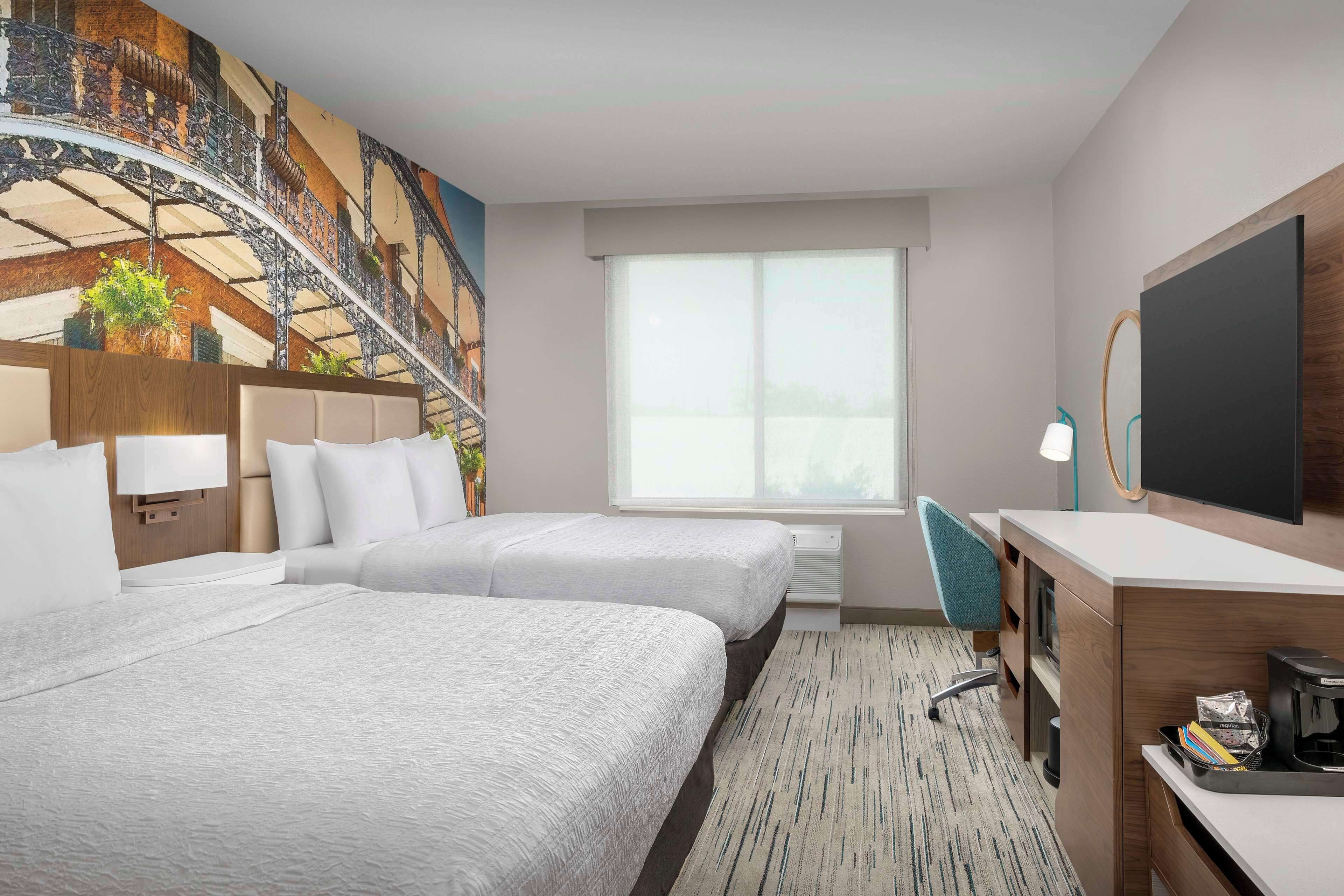Ein Hotelzimmer mit zwei Betten, einem Schreibtisch, einem Stuhl, einem großen Fenster und einem Wandgemälde.