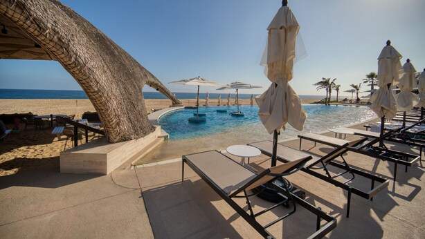 Un espace piscine aménagé au bord de la plage, avec des chaises longues et des parasols, une structure en chaume et une vue dégagée sur l’océan.