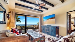 Un salon spacieux doté d’une grande fenêtre donnant sur une plage, d’un ventilateur de plafond et d’un téléviseur fixé au mur.